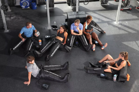 A group of athletes rests in a gym after a workout and uses NormaTec boots to aid recovery