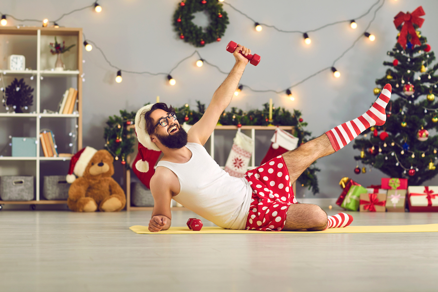 Happy beginner athlete in funny underwear and Santa hat made New Year resolution to keep fit and healthy and is doing supine side leg raises with dumbbells during home workout on Christmas holidays
