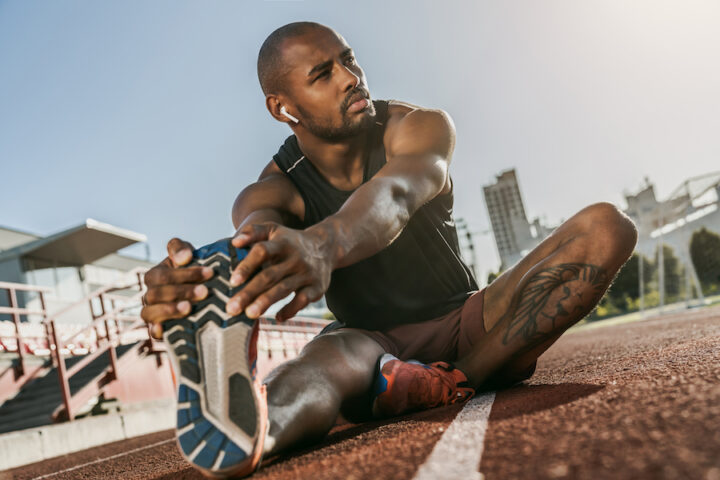 Sporty muscular african male athlete in earphones looking away, stretching legs while sitting at the stadium race track