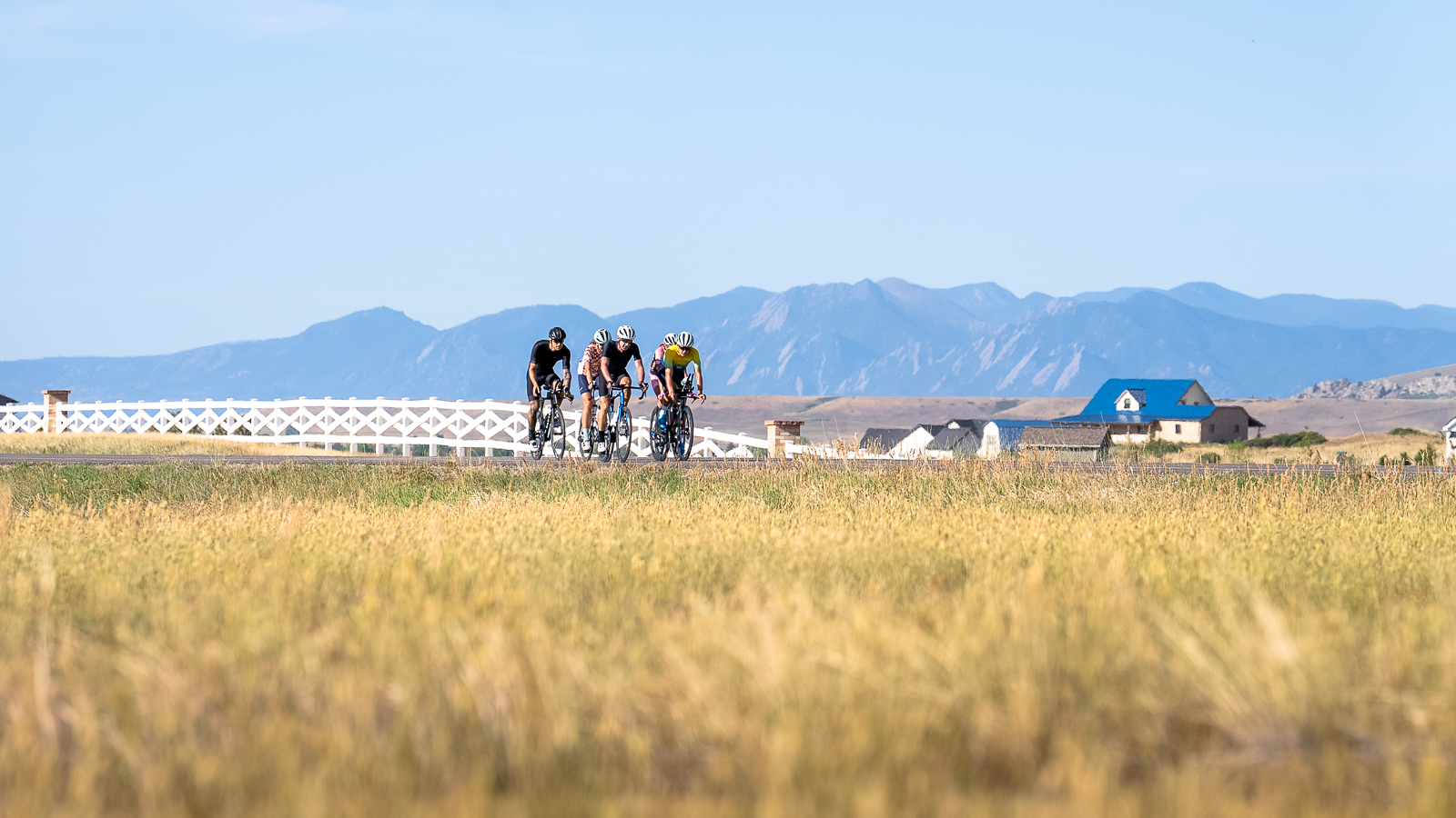 A group of strong cyclists affiliated with Fast Talk Labs rides along a fence with a farm and mountains in the background near Boulder, Colorado.