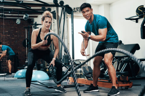 A woman exercising with battle ropes while her coach encourages her