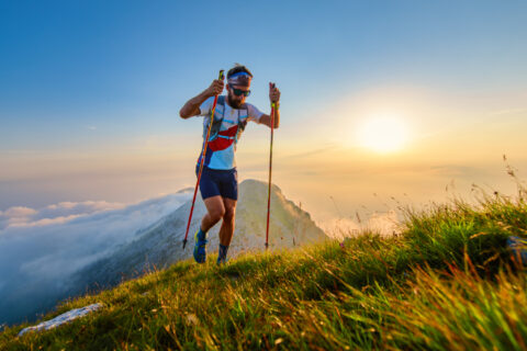 An ultra-runner summits a mountain peak with the sun low in the sky behind him