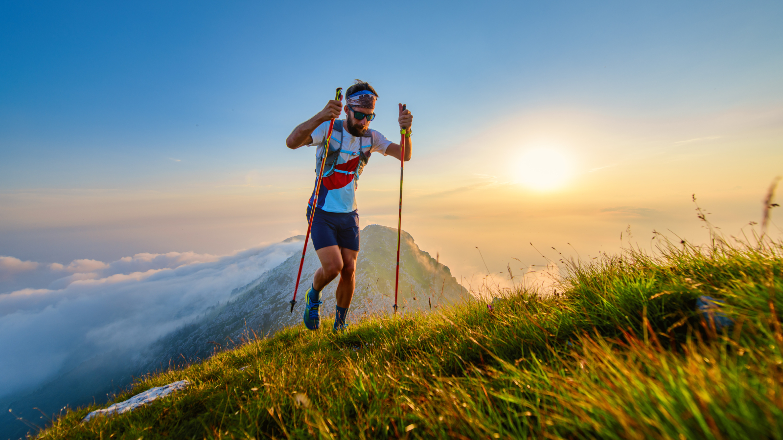 An ultra-runner summits a mountain peak with the sun low in the sky behind him