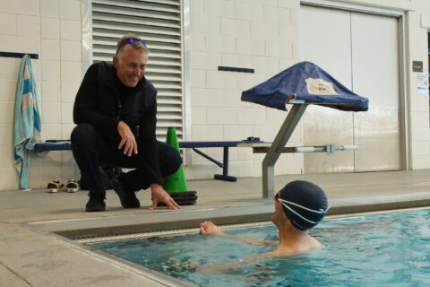 Joe Friel kneels at the edge of an indoor pool to talk with an athlete in the water
