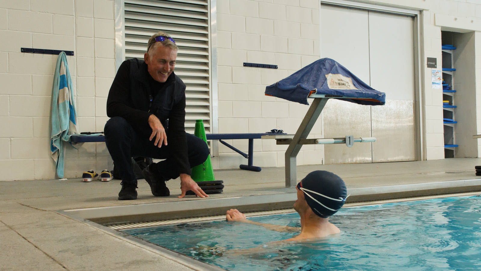 Joe Friel kneels at the edge of an indoor pool to talk with an athlete in the water