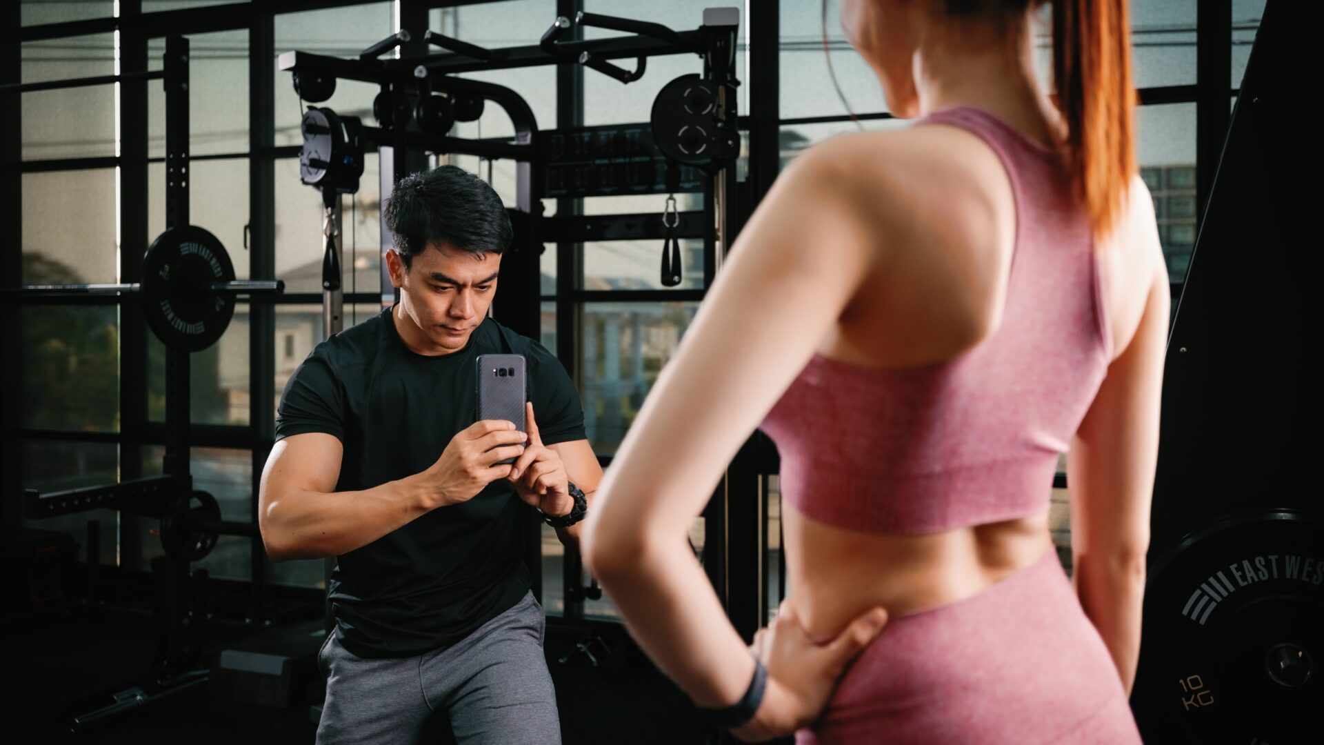 Man taking a photo with his phone of a female fitness influencer at the gym.