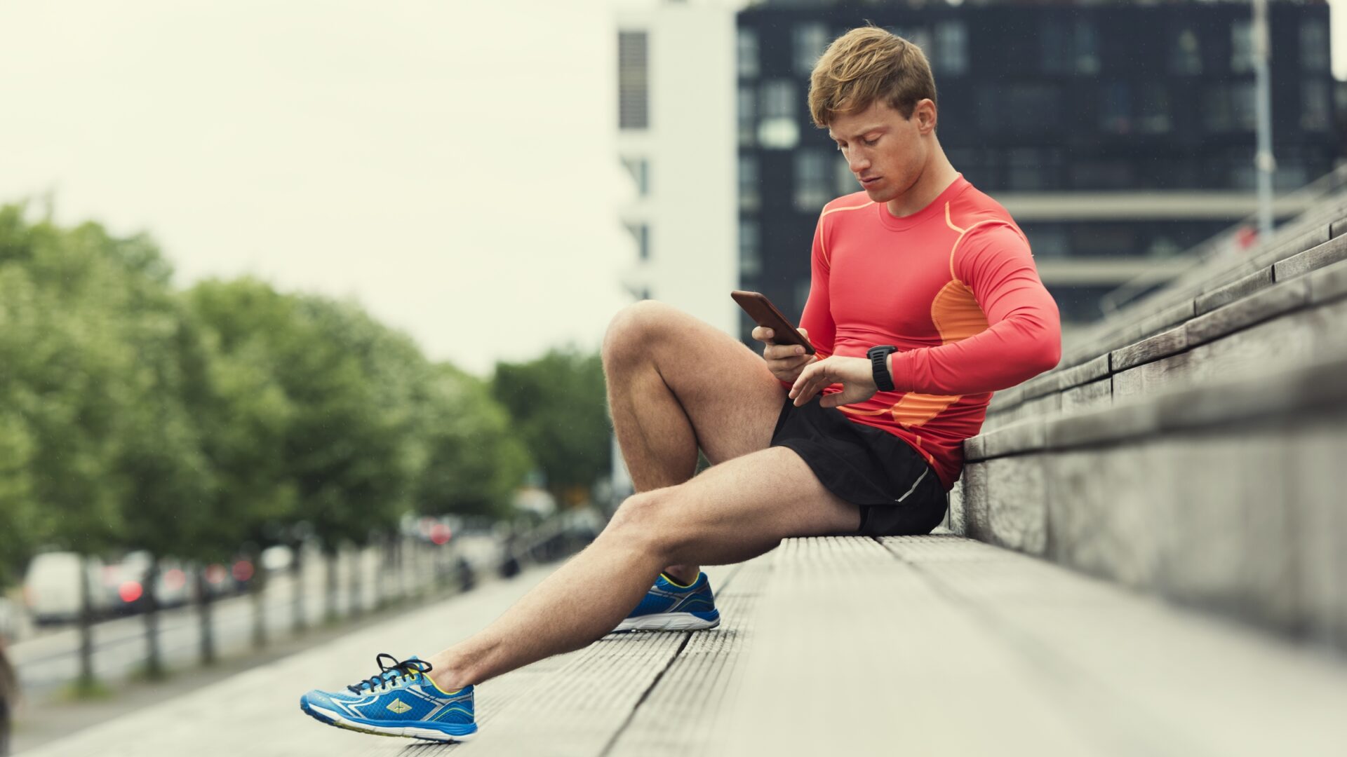 A runner rests on concrete steps while checking data on their watch and phone.