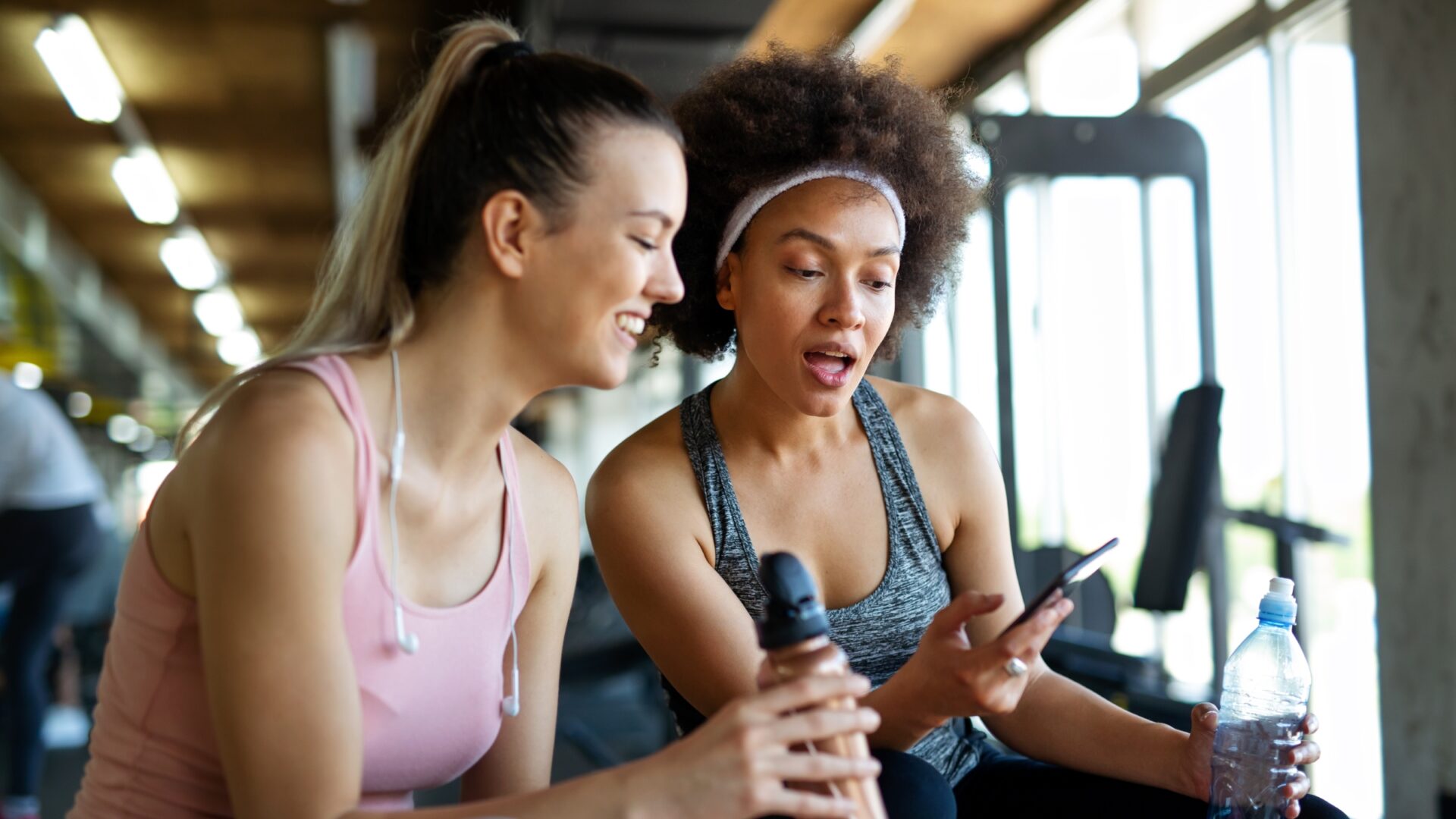 Two women at the gym smile at something on a phone.