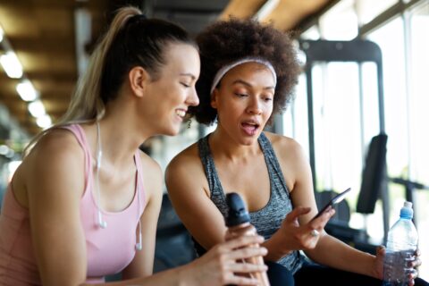 Two women at the gym smile at something on a phone.