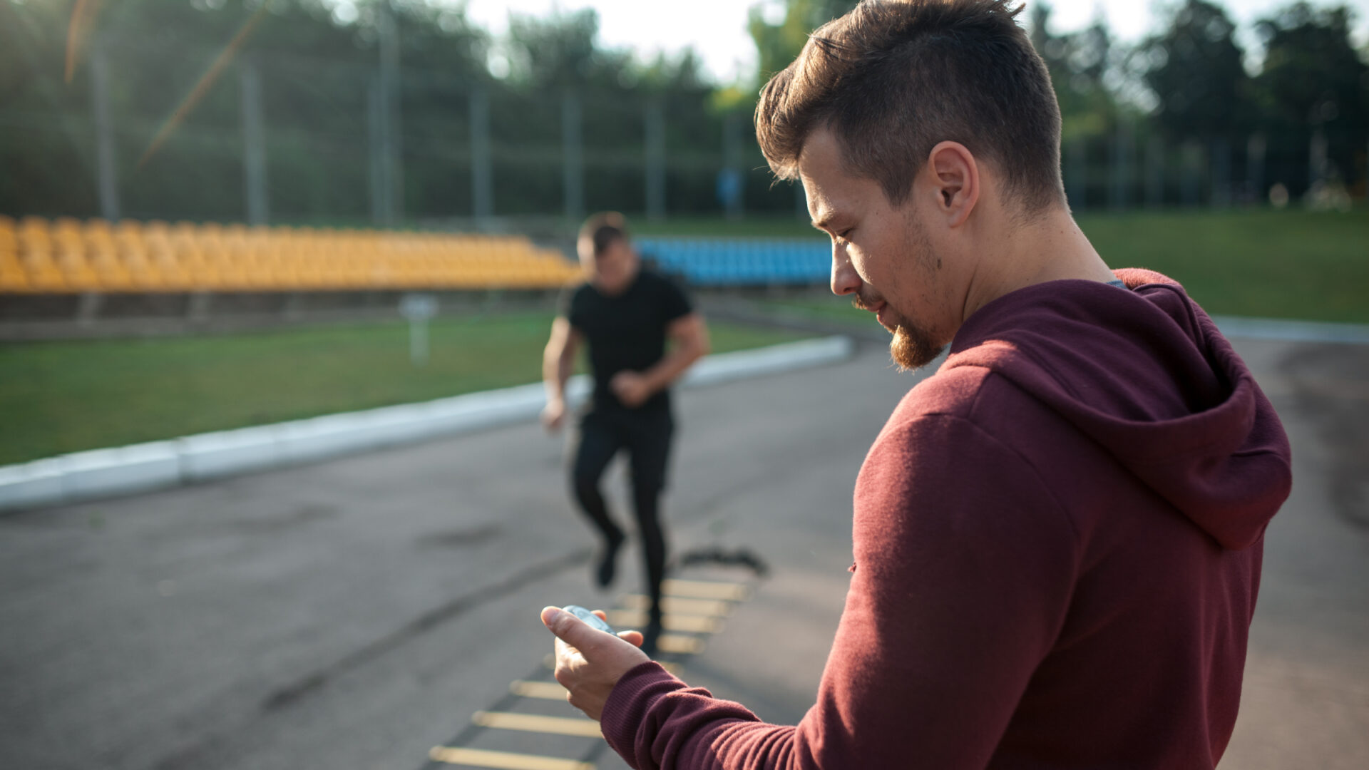 A coach looks at their stopwatch while their athlete does a ladder exercise in the background