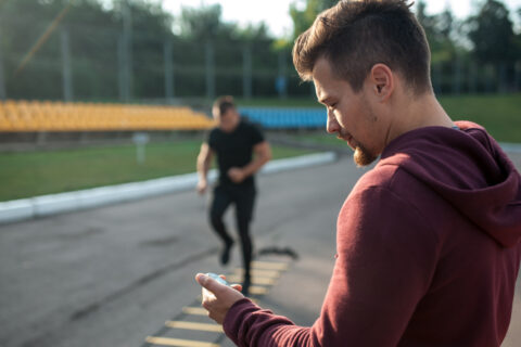 A coach looks at their stopwatch while their athlete does a ladder exercise in the background