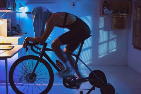 Cyclist rides on an indoor trainer in a dark room. He has his shirt covering his head