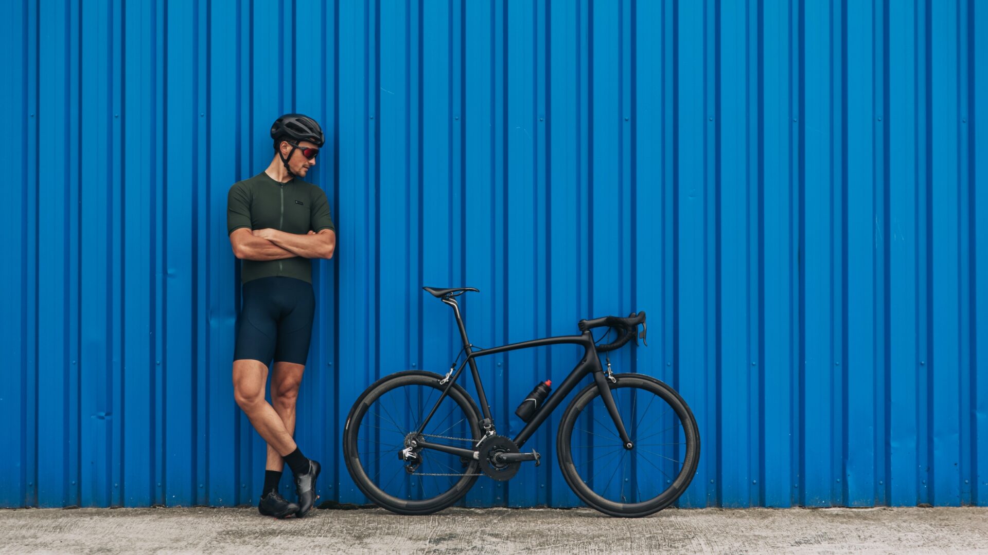 Man leans against a blue shipping container, looking at his all-black bicycle