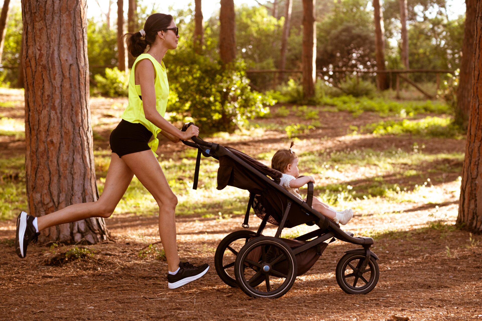 Woman running through a forest while pushing a baby stroller, returning to sport after giving birth.